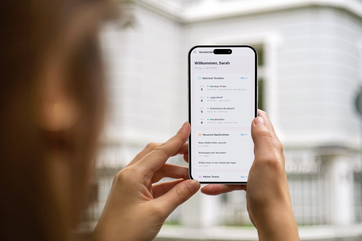Person hält Smartphone mit der Qehillah Gemeinde-App vor einem hellen Gebäude